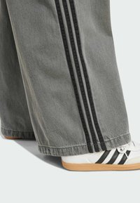 Pantalones de mezclilla de pierna ancha en gris con rayas negras en los lados. Una zapatilla blanca con rayas negras y una suela de goma es parcialmente visible.
