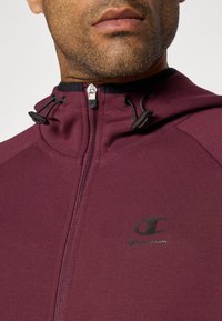Nahaufnahme des unteren Gesichts und des Halses eines Mannes, der einen weinrotfarbenen Reißverschluss-Hoodie mit schwarzen Kordeln und einem Champion-Logo auf der Brust trägt.