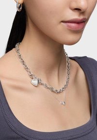 Collar de cadena de plata con un colgante de corazón brillante, en capas con una cadena delicada que sostiene dos pequeños acentos de corazón. Textura lisa.