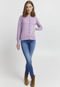 Cardigan de culoare violet deschis, cu guler rotund, închidere frontală cu butoni și manșete rasucite. Purtați cu blugi skinny albaștri și cizme scurte maronii.
