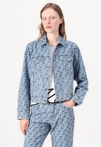 Chaqueta de denim azul claro con un patrón geométrico en toda la superficie, dos bolsillos en el pecho, cierre de botones y un ajuste ligeramente corto.