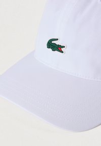 Λευκό καπέλο μπέιζμπολ με υφή υφάσματος, κυρτή γείσο και πράσινο λογότυπο κροκοδείλου με κόκκινες λεπτομέ ρειες μπροστά.