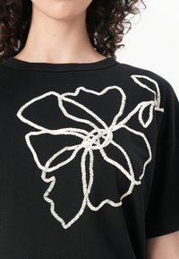 T-shirt noir avec un grand motif de fleur brodé blanc. Col rond, manches courtes, texture lisse et accents de couture détaillés.