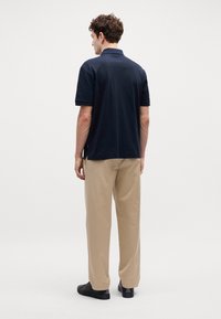 Polo de marinha com mangas curtas, textura lisa; combinado com calças bege claras; sapatos pretos; etiqueta da marca visível na bainha da camisa.