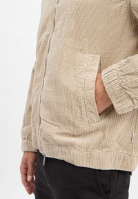 Beige corduroy jas met ribbeltextuur, ritssluiting en zijzakken. Manchetten en zoom zijn elastisch. Gecombineerd met zwarte broek.