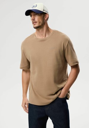 Mężczyzna w beżowej koszuli z krótkim rękawem i niebieskiej czapce z daszkiem, stoi z lewą ręką w kieszeni i patrzy w bok.