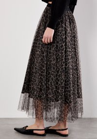Frau in einem braun-schwarzen Leopardenmuster-Tüllrock, schwarzem cropped Sweater und schwarzen Slingback-Ballerinas, die auf einem grauen Boden steht.