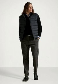 Άνδρας φορεμένος με μαύρη γιλέκα πάνω από μαύρο πουλόβερ και ανοιχτό μπλε πουκάμισο, γκρι παντελόνι και μαύρα επίσημα παπούτσια, στέκεται με τα χέρια στις τσέπες.