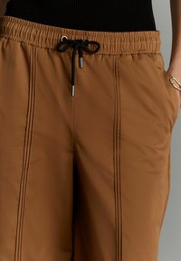 Pantaloni maro cu talie elastică și șnur negru. Au două buzunare laterale și cusături verticale care se întind pe partea din față pentru un plus de detaliu.
