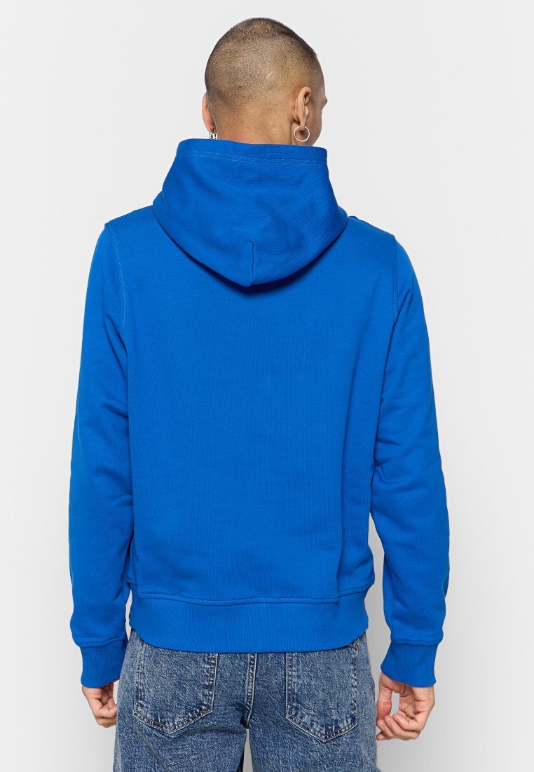 Persona con cabello corto y pendientes, vistiendo un sudadera azul brillante y jeans azules deslavados, vista desde atrás contra un fondo liso.