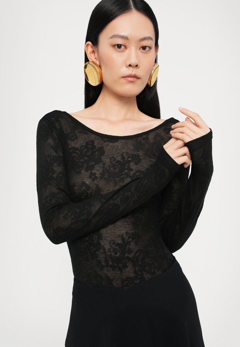 Body in pizzo nero con maniche lunghe, motivo floreale, scollo a pupazzo e silhouette aderente, abbinato a grandi orecchini circolari dorati.