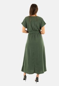 Vestido maxi verde con mangas cortas, cintura con cinturón y tejido fluido. Textura suave, diseño hasta el suelo, silueta simple y detalle de abertura.