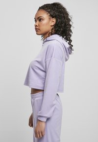 Ungdommelig kvinne med krøllete hår iført en lavendel cropped hettegenser og matchende joggebukser, stående i profil mot en ensfarget bakgrunn.