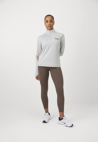 Lys grå zip-up sportstrøje med lange ærmer og Nike-logo, parret med tætsiddende brune leggings og hvide sneakers med marineblå detaljer.
