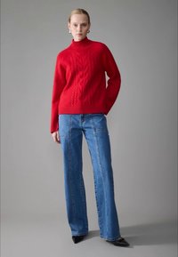 Suéter de gola alta em malha canelada vermelha, combinado com jeans de perna larga azul claro, com costuras frontais. Sapatos pretos de bico fino completam o look.