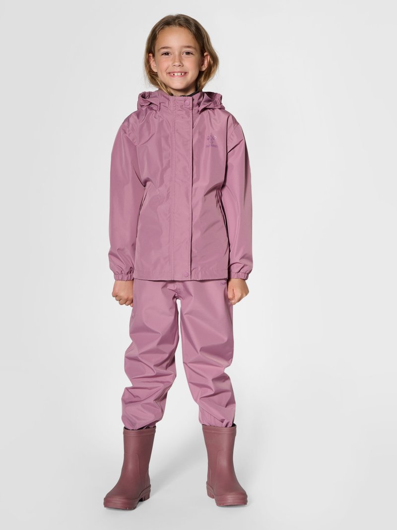 Junges Kind trägt eine passende pinke Regenjacke, Hose und Gummistiefel, lächelt und steht auf einem einfarbigen Hintergrund.