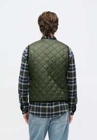 Άνδρας με πλάτη, φορώντας ένα πράσινο καπιτονέ γιλέκο πάνω από ένα μπλε και πράσινο καρό πουκάμισο με ανοιχτό μπλε τζιν, μπροστά σε απλή φόντο.