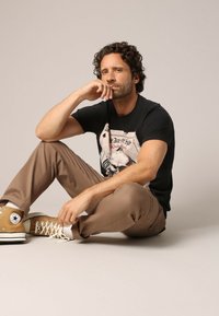 T-shirt noir avec un design graphique, chinos beiges et baskets montantes marron clair. Homme assis, avec des cheveux bouclés, la main sur le menton.