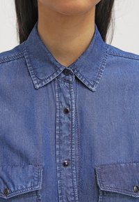 Modrá džínová košile s špičatým límcem, viditelným šitím, předním zapínáním na knoflíky a dvěma náprsními kapsami. Hladká textura, neformální design.