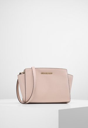 Petit sac à épaule en cuir rose blush Michael Kors avec une fine bandoulière et un logo doré à l'avant, présenté sur une surface blanche.