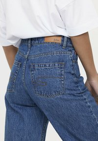 Jeans de mezclilla azul con una cintura alta, que cuentan con dos bolsillos traseros y costuras amarillas en contraste. La etiqueta de cuero en la cintura dice "Le Cooper."