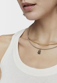 Tres collares: un collar choker de metal dorado y plateado, con una cadena plateada que presenta un pequeño charm negro en forma de oso.