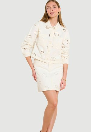 Mladá žena oblečená v bielom gombíkovom saku a sukni s kvetinovými výrezmi, stojaca na jednoduchom svetlom pozadí.