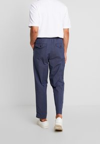 Man med marinblå byxor med knäppta bakfickor och vita sneakers, stående mot en enkel ljus bakgrund.