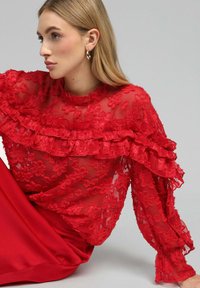 Blouse en dentelle rouge avec des détails froncés et des manches longues. Un motif floral transparent recouvre une base rouge unie. Texture lisse avec une légère transparence.