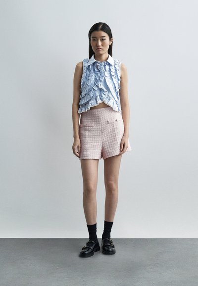 Νέα γυναίκα που φορά αμάνικη μπλούζα με μπλε ρίγες και βολάν, ροζ σορτς με υφή, μαύρες κάλτσες και μαύρα μοκασίνια, στέκεται μπροστά σε έναν απλό τοίχο.