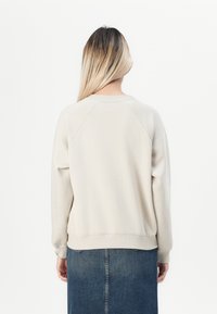 Sudadera beige claro con mangas raglán, cuello redondo y puños acanalados. Lleva una falda de mezclilla, mostrando un diseño simple y casual.