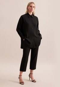 Schwarzes Hemd mit Knopfleiste, entspannter Passform und langen Ärmeln, kombiniert mit cropped schwarzen Hosen und schwarzen Riemchen-Highheels. Glatte Textur.