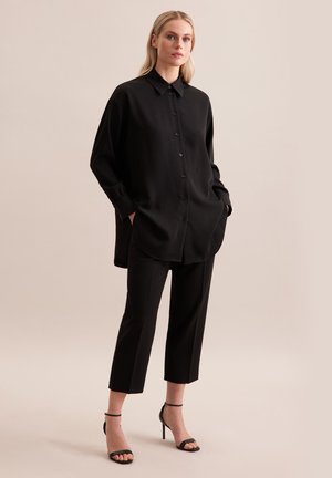 Camicia nera a maniche lunghe con un taglio rilassato, abbinata a pantaloni neri a tre quarti e tacchi neri con strappi. Texture liscia.