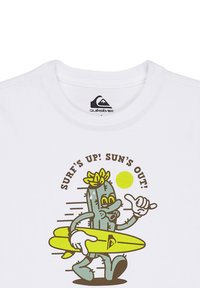 Bílý bavlněný tričko s karikovým kaktusem, který drží žlutou surfovou desku, a nápisem "SURF'S UP! SUN'S OUT!" hnědou barvou.