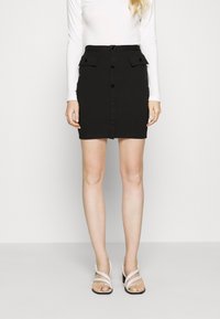 Zwarte, aansluitende rok met een knoopsluiting aan de voorkant, twee klepzakken en een gladde textuur, gecombineerd met een witte blouse met lange mouwen en lichtgekleurde hakken.