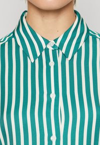 Teal ja valge triibuline nööbideta särk klassikalise kraega, millel on viis valget nööpi ees, silmapaistva sileda kangastekstuuri jaotusega.