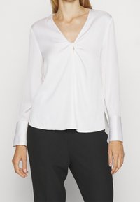 Bílá saténová blůza s dlouhým rukávem a véčkovým výstřihem, s předním twist detailem a kontrastními lesklými manžetami; volný střih a hladká textura.