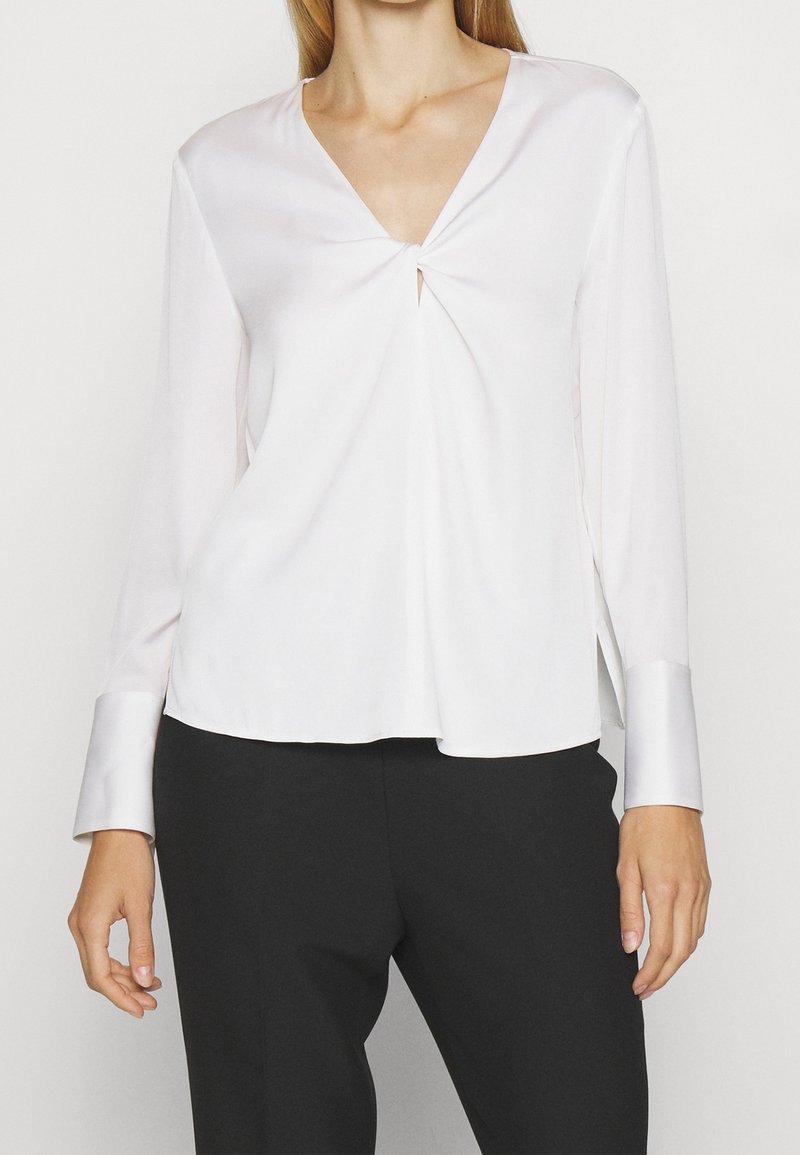 Bílá saténová blůza s dlouhým rukávem a véčkovým výstřihem, s předním twist detailem a kontrastními lesklými manžetami; volný střih a hladká textura.