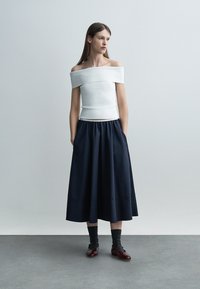 Vrouw met een wit off-shoulder top, een marineblauwe midi-rok met zakken, donkere sokken en bordeauxrode Mary Jane schoenen, staand tegen een grijze achtergrond.