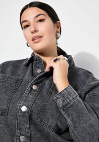 Chaqueta de mezclilla gris con botones de presión plateados, un cuello y grandes bolsillos. El modelo lleva un delineador de ojos naranja intenso y varios anillos. Textura suave.