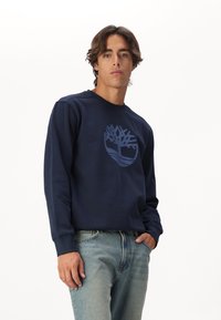 Námořnická crewneck mikina vyrobená z měkké látky s světle modrým logem stromu. Nosit s světle modrými džíny, což ukazuje na ležérní styl.
