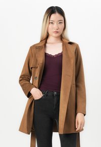 Ung kvinne med rett svart-blondt hår, iført en brun frakk, mørk lilla bluse med blondekanter og svarte jeans, står mot en hvit bakgrunn.