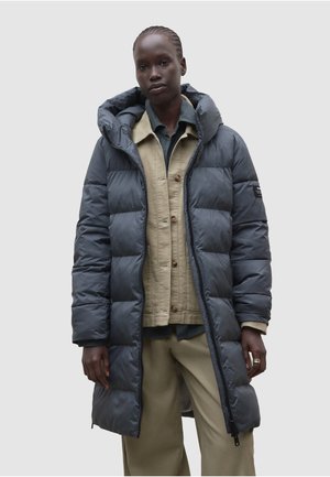 Persona alta que lleva un abrigo acolchado largo de color gris oscuro sobre una chaqueta beige abotonada y pantalones beige sobre un fondo liso.