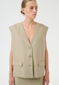 Beige bez Piedurkņu veste ar dziļu V-veida kakla izgriezumu, divām priekšējām kabatām un trīs pogu aizdari. Izgatavota no viegla auduma ar gludu tekstūru.