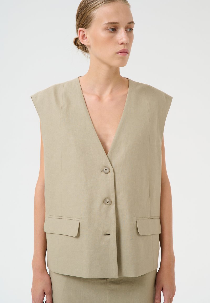 Beige bez Piedurkņu veste ar dziļu V-veida kakla izgriezumu, divām priekšējām kabatām un trīs pogu aizdari. Izgatavota no viegla auduma ar gludu tekstūru.