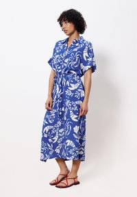 Blaues Kleid mit weißen Blumenmustern, kurzen Ärmeln, einer Knopfleiste vorne und einem gebundenen Taillengürtel, kombiniert mit roten Riemchensandalen.