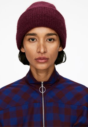 Gorro de punto burdeos con puño doblado; la modelo lleva una chaqueta con cremallera en un patrón de cuadros púrpura y azul, con un alto cuello.