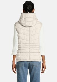 Vrouw met donker haar in een bun, gekleed in een mouwloze beige gewatteerde puffervest met capuchon over een witte geribbelde langemouwen top en blauwe jeans.