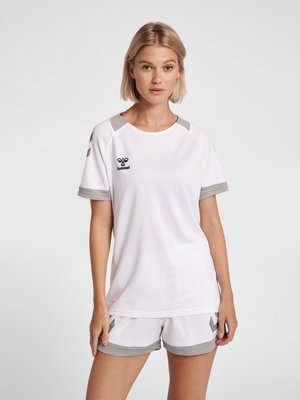 Blonde vrouw draagt een wit-grijs Hummel sportoutfit met t-shirt en korte broek, staand in ontspannen houding tegen een effen achtergrond.