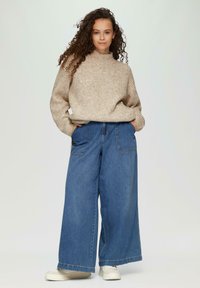 Beige gestrickter Pullover mit hohem Kragen, kombiniert mit weit geschnittenen blauen Jeans aus Denim mit Fronttaschen. Weiße Schuhe runden den Look ab.